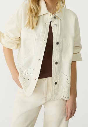 Vrouw draagt crèmekleurig jasje met bloemcut-out details over een bruin topje en crèmekleurige broek, mouwen opgerold, hand in zak.