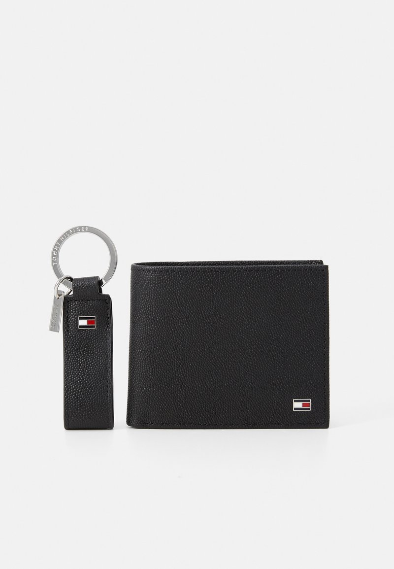 Portefeuille en cuir noir avec une texture et un porte-clés. Présente un petit emblème et un anneau de clé attaché par une sangle. Design compact.