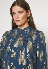 Μπλε μπλούζα με χρυσό γυαλιστερό σχέδιο ζώου, πλισέ σχέδιο, ψηλό γιακά με κουμπί κλεισίματος και φουσκωτά μανίκια με υφή.