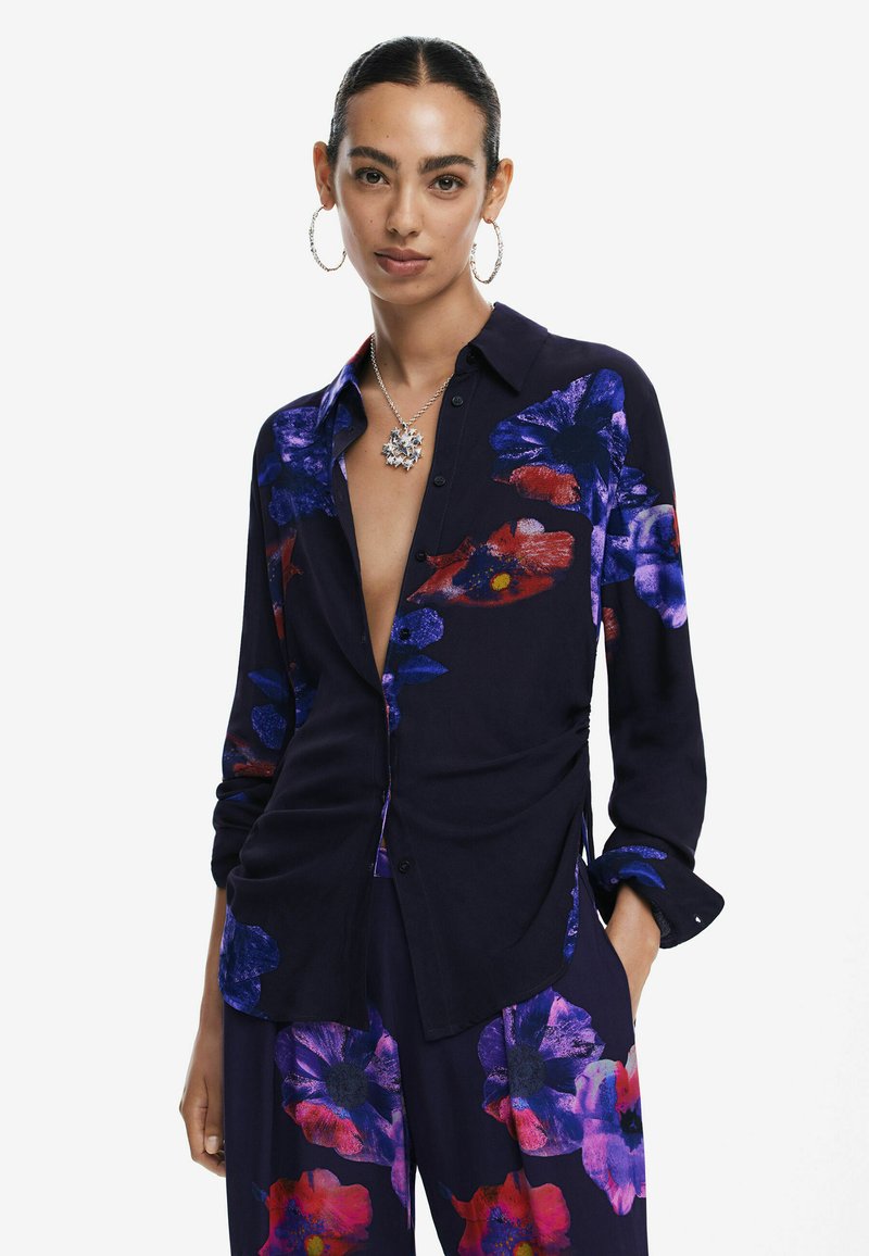Marinebluse mit großen lila und roten Blumenmustern, langen Ärmeln, Knopfverschluss und seitlichen Schlitzen. Das Modell trägt silbernen Schmuck.