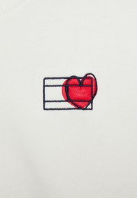 La tela blanca presenta un diseño bordado de un corazón rojo parcialmente encerrado en un contorno negro que recuerda a un marco rectangular.