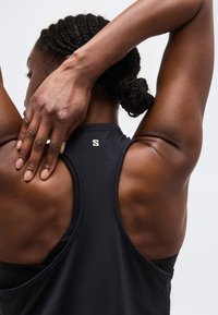 Žena s vrkočmi vo vlasoch, oblečená v čiernom športovom tielku bez rukávov, natiahne pravú ruku za hlavu na jednoliatom pozadí.