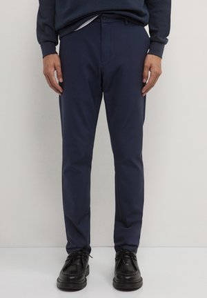 Marineblaue Hose aus glattem Stoff, Slim-Fit-Design und mit Fronttaschen. Kombiniert mit schwarzen Schuhen und dezenten Details.