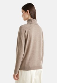 Žena s dlouhými hnědými vlasy, oblečená v béžovém roláku a bílých kalhotách, stojící bokem před neutrálním pozadím.