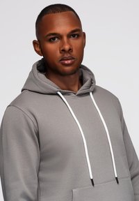 Jeune homme aux cheveux courts portant un sweat à capuche gris avec des cordons blancs, regardant droit devant sur un fond clair et uni.