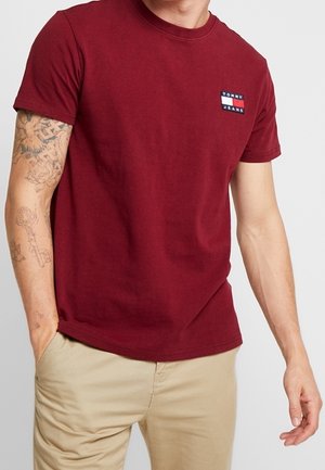 Burgundowa bawełniana koszulka z krótkim rękawem, z małym niebiesko-białym-czerwonym prostokątnym logo na lewej piersi. Prosty design, o okrągłym dekolcie.