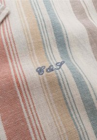 Tessuto morbido con righe orizzontali in rosso tenue, beige, blu e grigio, caratterizzato da piccole lettere ricamate "C & S" in filo blu.