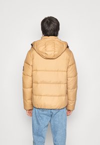 Pufjakke i lys tan med et isoleret design, der har hætte og syede paneler. Bæres over blå jeans.