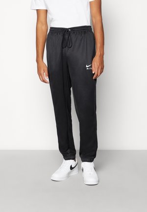 Man draagt zwarte Nike Air trainingsbroek met trekkoord en witte Nike sneakers tegen een witte achtergrond.