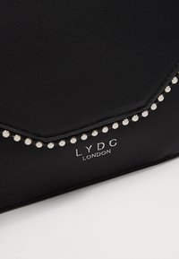 Borsa in finta pelle nera con borchie argento lungo la patta, dotata di un logo prominente "LYDC LONDON" in lettere metalliche.