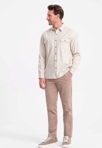 Lichtbeige overhemd met knopen en twee borstzakken, gecombineerd met lichtbruine corduroy broek en beige sneakers met witte zolen.