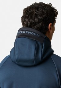 Felpa blu navy con colletto alto e rifiniture nere. Presenta la scritta "SWEDEMOUNT" in testo grigio sul colletto. Tessuto liscio e strutturato.