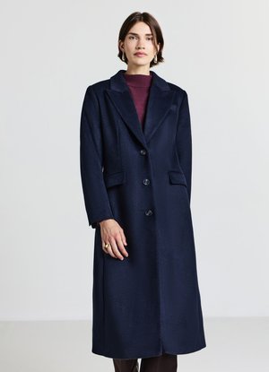 Donna con un lungo cappotto blu navy con bottoni sopra una maglia bordeaux, in piedi davanti a uno sfondo chiaro uniforme.