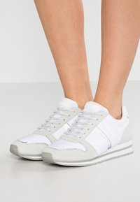 Baskets blanches et grises avec une finition en cuir lisse et en daim, dotées d'une semelle texturée, de rayures contrastantes et d'œillets métalliques pour les lacets.