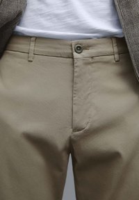 Beige chinos med en slät yta, som har knäppning och fickor framtill utan veck. Diskreta sömdetaljer nära midjan.