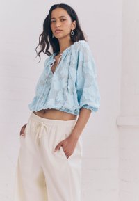 Top corto azzurro chiaro con texture e scollo arricciato, abbinato a pantaloni crema larghi con vita regolabile a coulisse e tasche laterali.