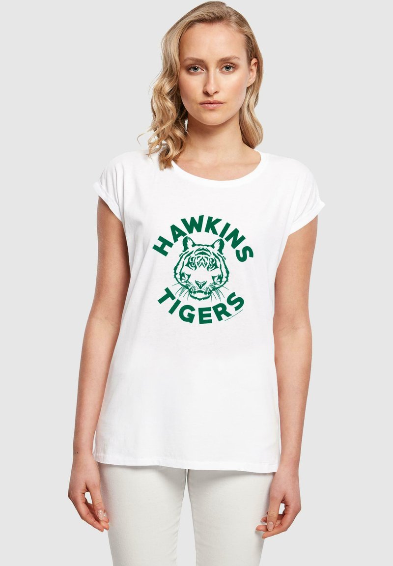 Baltas kokvilnas T-krekls ar izvirpotām piedurknēm, ar zaļu grafiku, kurā attēlots tigers un uzraksts "HAWKINS TIGERS" treknajā burtu izpildījumā.