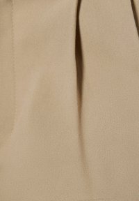Beżowy, teksturowany materiał o gładkim wykończeniu, z delikatnymi fałdami i subtelnymi detalami przeszycia, świadczący o lekkim składzie.