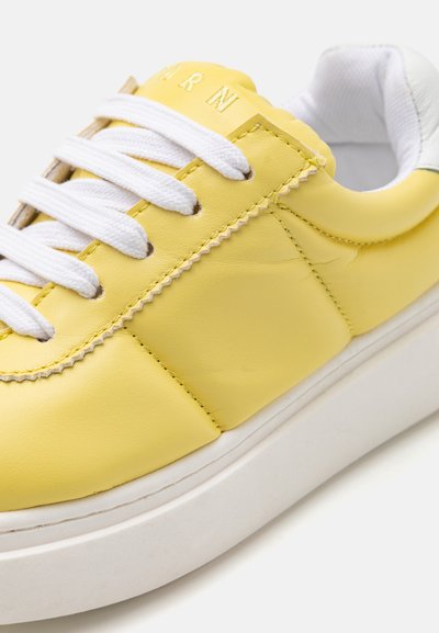 Gul lädersneaker med vita snören, vadderad krage och en strukturerad yta. Har en tjock vit sula och dekorativ sömnad.