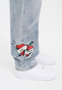 Lyseblå denimjeans med broderede røde hjerter og et bånd med "Ed Hardy" tekst på venstre manchet, båret med hvide sneakers.