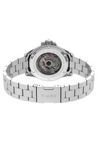 Rozsdamentes acél óra ezüst szíjjal, kerek tok a hátoldalán átlátszó részel, ami a szerkezetet mutatja, "TIMEX" és "WATER RESISTANT 50M" felirattal.