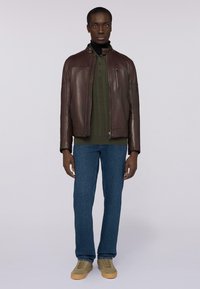 Bruine leren jas met staande kraag, gecombineerd met een groene ribbeltrui, blauwe jeans en grijze sneakers. Model staat tegen een lichte achtergrond.