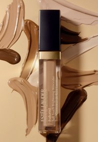 Corrector iluminador de textura suave Futurist de Estée Lauder en un tubo transparente con tapa negra y detalles en dorado, rodeado de diversas muestras de cremas en tonos beige y marrón.