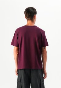 Mann, der von hinten schaut, trägt ein schlichtes, dunkelviolettes Kurzarm-T-Shirt und schwarze Cargo-Hosen, steht vor einem weißen Hintergrund.