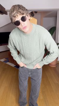 Világos zöld, kötött pulóver fonott mintával; szürke, bő szárú nadrág; kerek fekete napszemüveg; beltéren, fából és növényekből álló háttérrel.