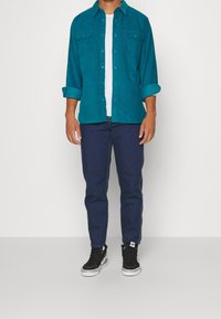 Camicia in velluto a coste teal con bottoni sul davanti e maniche arrotolate; t-shirt bianca sotto; jeans blu scuro; sneakers nere con dettagli bianchi.