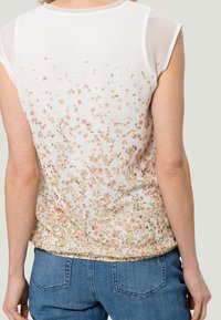 Mujer que lleva una blusa blanca con pequeño estampado floral en rosa y verde y mangas de red, combinada con jeans azul.