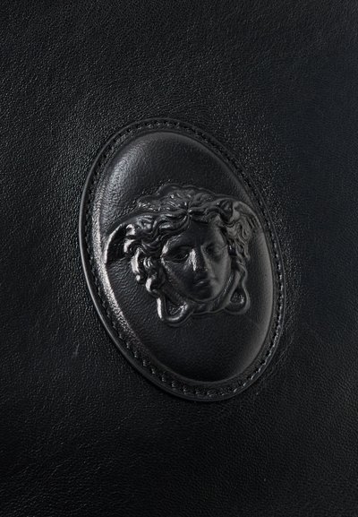 Μαύρη δερμάτινη επιφάνεια με ανάγλυφο οβάλ έμβλημα που απεικονίζει λεπτομερή κεφαλή της Μέδουσας στο κέντρο.