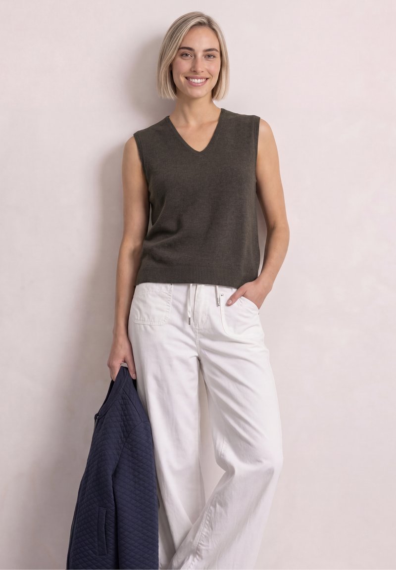 Femme aux cheveux blonds courts portant un haut sans manches foncé et un pantalon blanc, tenant une veste matelassée bleu marine dans une main.