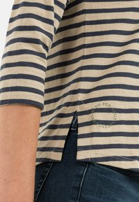 Beige and navy striped long-sleeve shirt with side slit worn over dark blue denim jeans, showing part of the arm and hip.