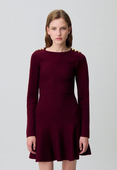 Vestido burdeos de manga larga con un corsé ajustado y una falda acampanada. Presenta detalles de botones dorados en el hombro. Tejido de punto suave.