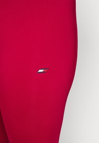 Leggings de sport rouges en tissu extensible présentant un petit logo bleu, blanc et rouge sur le côté gauche. Texture lisse et design ajusté.