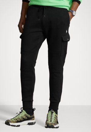 Pantalon jogging cargo noir avec poches latérales, porté avec des baskets vertes et camouflage, et un sweat vert partiellement visible.