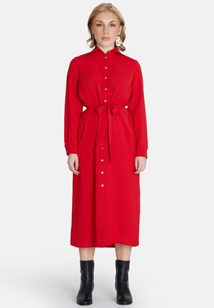 Rotes, langärmeliges Kleid mit einem Kragen, vorderen Knöpfen und einer gebundenen Taille. Die Länge reicht unter das Knie und wird mit schwarzen Knöchelstiefeln kombiniert.