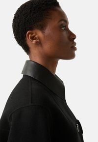 Sort frakke med stor læderkrave, struktureret silhuet og tekstureret stof. Knapdetalje synlig foran.