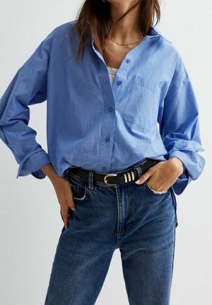Frau trägt ein hellblaues Hemd mit Knöpfen, das in eine hoch taillierte dunkelblaue Jeans mit schwarzem Gürtel gesteckt ist, steht mit den Händen in den Taschen.