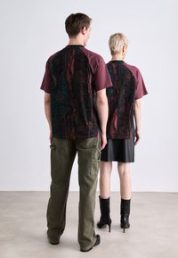 Twee personen dragen gecoördineerde korte mouwen shirts met een bordeauxrode voorkant en een donkere bedrukt achterzijde, gecombineerd met olijfgroene cargobroeken en een zwarte rok.