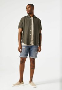 Olijfgroene korte mouwen shirt, beige rondhals t-shirt, versleten blauwe spijkerbroek shorts, tan instappers, met zichtbare tatoeages op het onderbeen.