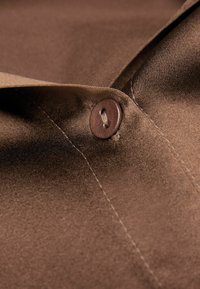 Bruine stof met een gladde textuur, voorzien van een bruine knop en zichtbare stikdetails. De stof lijkt iets glanzend.