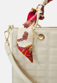 Cream-colored handbag with a quilted texture, featuring a gold ring, detachable strap, and a patterned silk scarf tied at the handle.