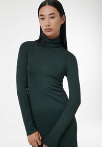 Robe verte à manches longues et col roulé avec une silhouette ajustée. Fabriquée dans un tissu texturé et scintillant qui capte la lumière, avec des détails subtils.