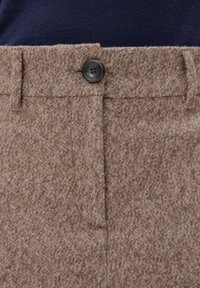 Pantalons en laine texturée marron avec un bouton noir et des passants de ceinture, portés avec un haut bleu marine foncé.