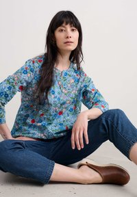 Blouse bleu à motifs floraux avec manches trois quarts, encolure ronde et accents rouges, associée à un jean en denim foncé et des sabots marron.