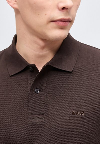 Homme portant un polo marron foncé avec un col et deux boutons, orné du logo "BOSS" sur la poitrine.