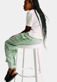 Witte t-shirt, groen en witte gestreepte broek en zwarte sandalen, gedragen terwijl ik op een eenvoudige witte kruk zit. Haar is in lange vlechten.