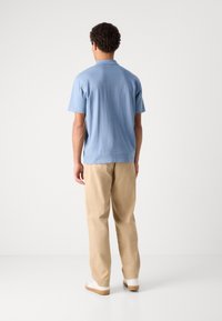 Cămașă polo cu mâneci scurte, de culoare albastru deschis, cu o croială lejeră și guler. Asortată cu pantaloni bej și adidași albi, văzută din spate.